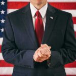 Prayer Twist Explodes In Iowa Chamber Man in suit with red tie standing before American flag