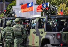 Beijing Tightens Noose – Chokehold Drills Rattle U.S. Soldiers beside camouflaged military vehicles with mounted guns and flags