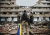 Soldier with rifle stands before a destroyed building holding a blue and yellow flag