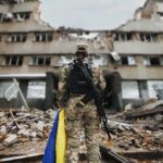 Soldier with rifle stands before a destroyed building holding a blue and yellow flag
