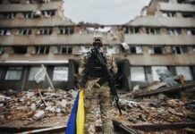 Deadly Kyiv Strikes Rattle Hospitals Soldier with rifle stands before a destroyed building holding a blue and yellow flag
