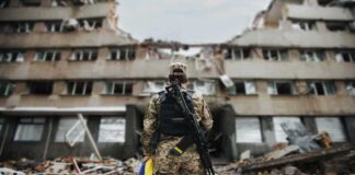 Soldier with rifle stands before a destroyed building holding a blue and yellow flag
